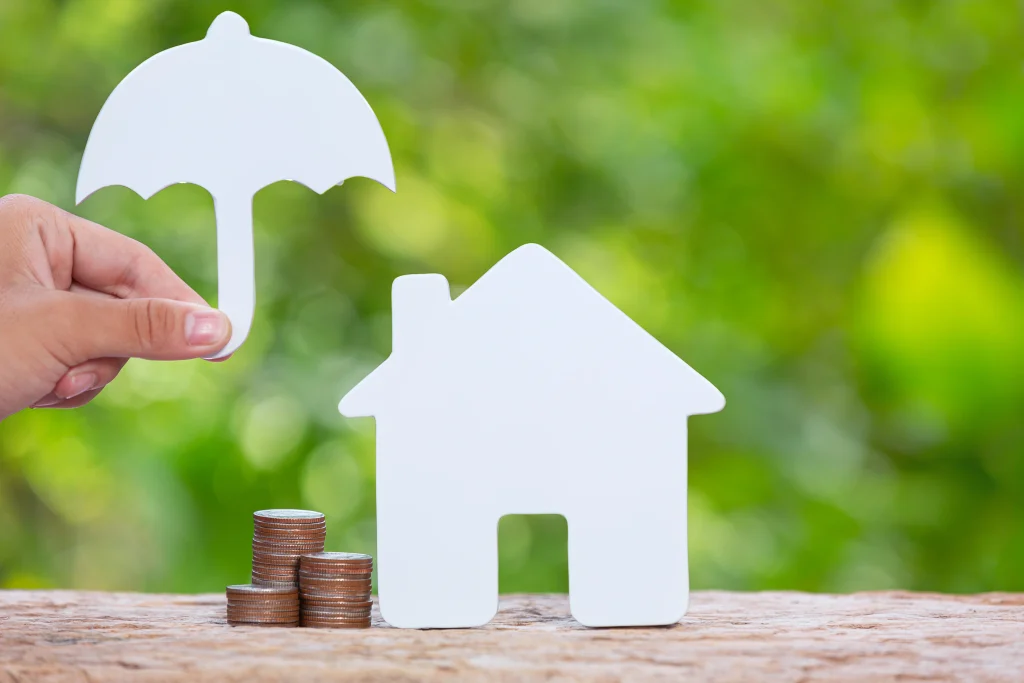 hand holding paper umbrella over house cutout and coin stacks home insurance