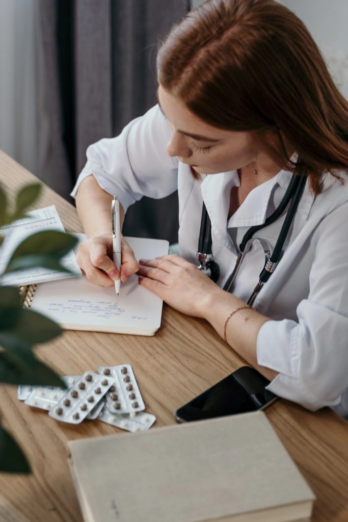 Healthcare professional writing medical notes at a desk with a stethoscope and medication nearby
